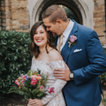 Elopement Wedding A bride in an off-shoulder white lace dress holds a vibrant bouquet as she smiles joyfully. A groom in a blue suit, white shirt, and rose boutonniere stands beside her, whispering into her ear. They are posed in front of a stone archway, with lush greenery surrounding them. Elopements Inc